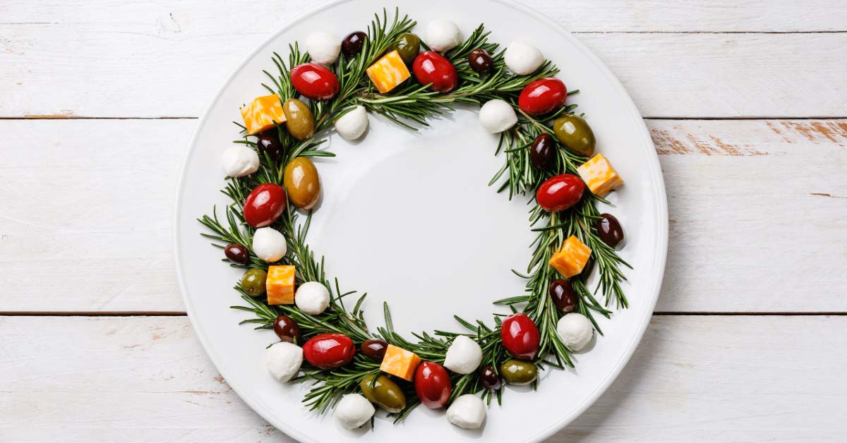 A wreath of rosemary sprigs with cheeses, tomatoes and olives on a white plate.