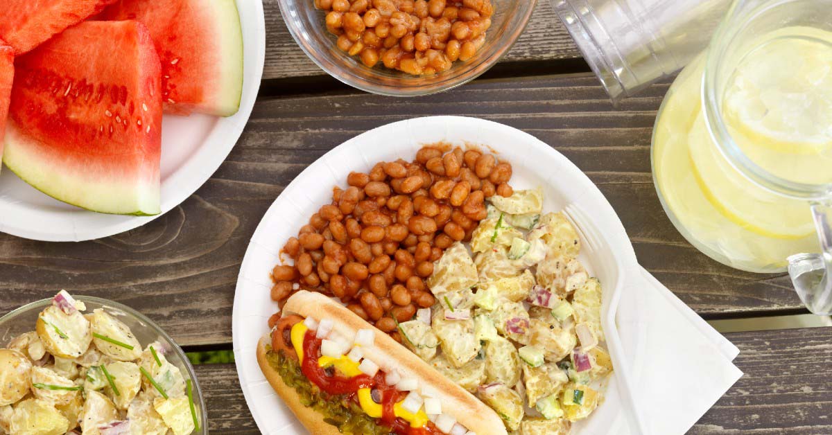 Picnic table setting with a plate of watermelon, bowls of baked beans and potato salad, a pitcher of lemonade and a plate with a brat, baked beans and potato salad.